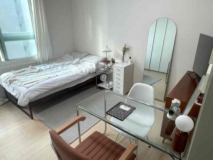 明るいナチュラルトーンのワンルーム寝室、ベッドと透明なガラスのダイニングテーブルが調和した雰囲気あるインテリア