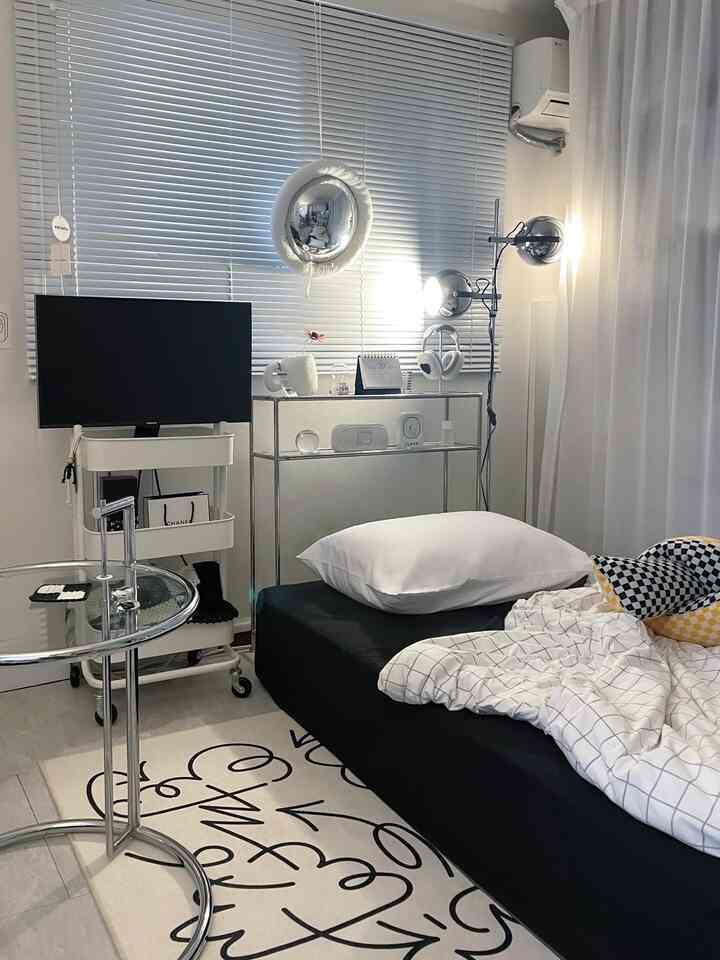 Modern single-bedroom interior with white and black tones, featuring a bed, metal side table, blinds, and curtains creating a cozy atmosphere