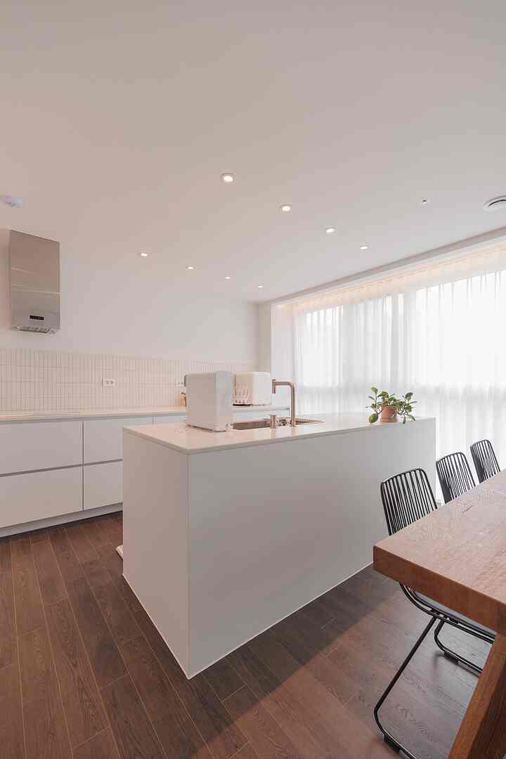 White and brown toned kitchen space featuring a central island, dining table on right, ceiling lights, and sheer curtains with a clean natural modern vibe