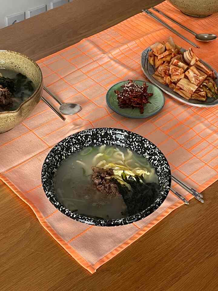 Warm kitchen dining table featuring enamel bowls with Korean soup dishes and kimchi on an orange table mat