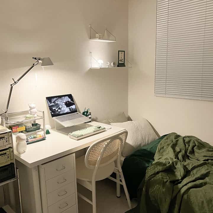 アイボリーとグリーンのトーンが調和したワンルームの書斎空間、デスク、椅子、壁棚、寝具が配置された落ち着いた雰囲気のインテリア