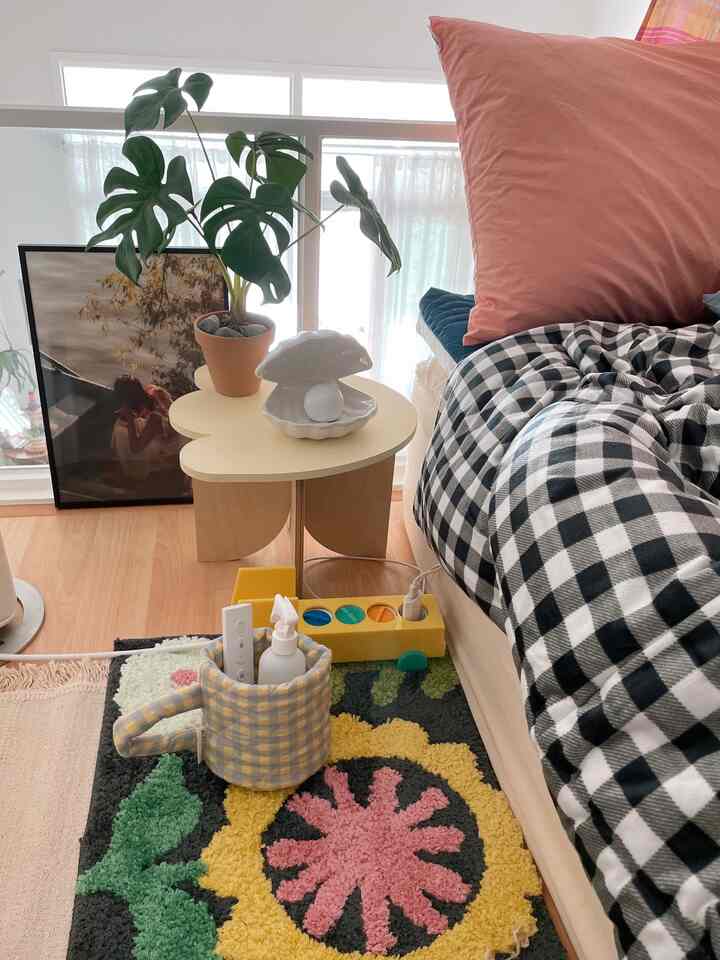 Cozy bedroom with white and wood tone background, checkered bedding, floral rug, flower-shaped side table, and potted plant