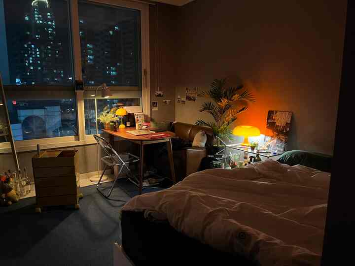 暖かいオレンジの照明と白い寝具があるワンルーム、デスクとブラウンのソファが配置された雰囲気のある空間