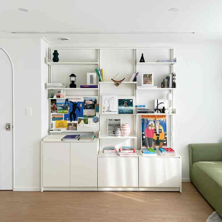 White-toned study space featuring clean system shelves with books and art, natural wood flooring, and a green sofa in a natural modern interior