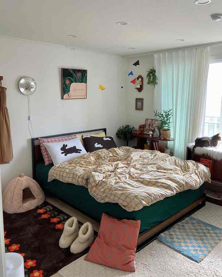 ナチュラルな木目調と白い背景の寝室、黄色のチェック柄の布団と黒いうさぎの枕が置かれた居心地の良い空間で、窓辺に猫が座っている