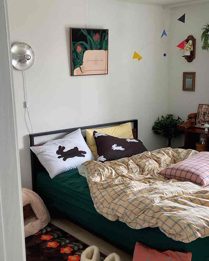 アイボリーの壁とダークグリーンのベッドがある自然でシンプルな雰囲気の寝室。黄色のチェックの掛け布団とウサギ柄の枕が特徴的なインテリア