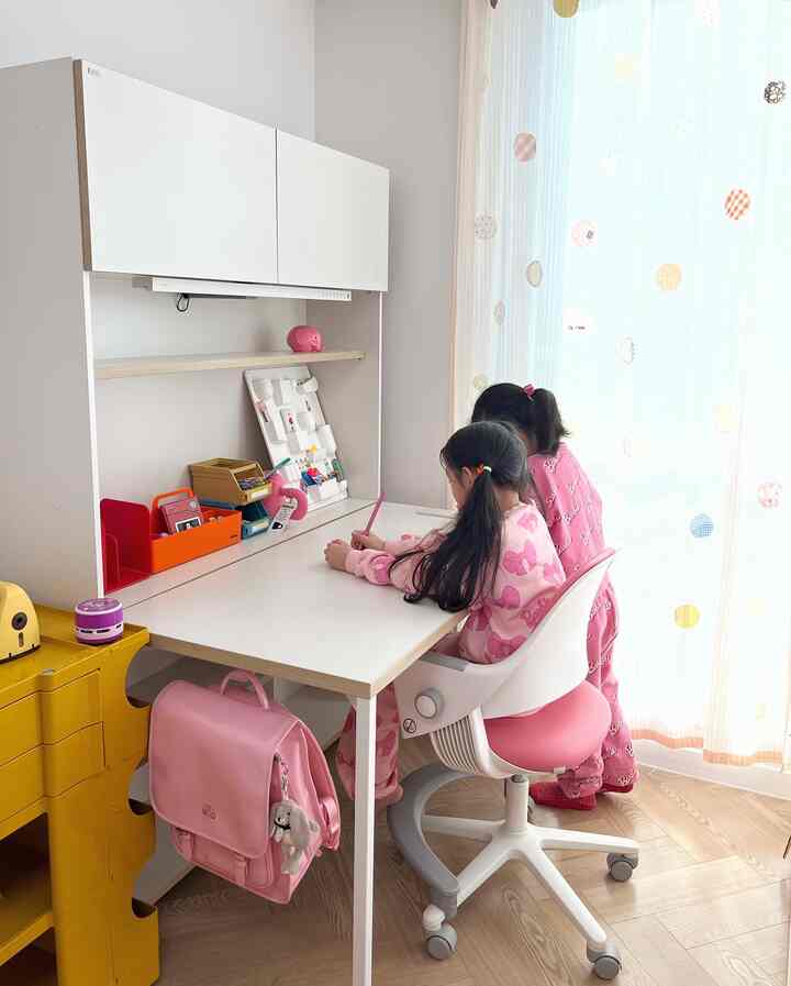 A white and pink toned kids' room with a desk and chair where children study, featuring natural wood flooring and bright curtains