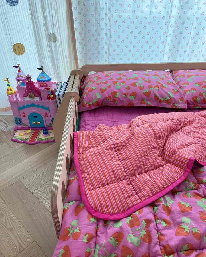 ピンクのいちご柄寝具が置かれた子供部屋のベッド、木目調のベッドフレームと白いカーテンが配置された印象的な空間