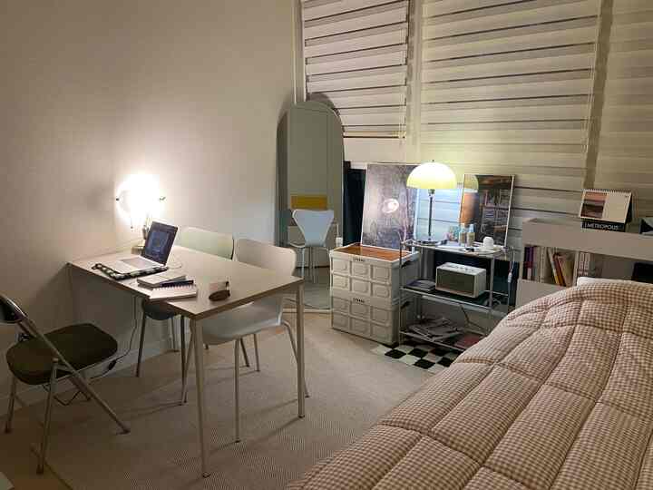 白とベージュのトーンのワンルーム、デスクとベッド、照明が配置された清潔でモダンな一人暮らしの空間