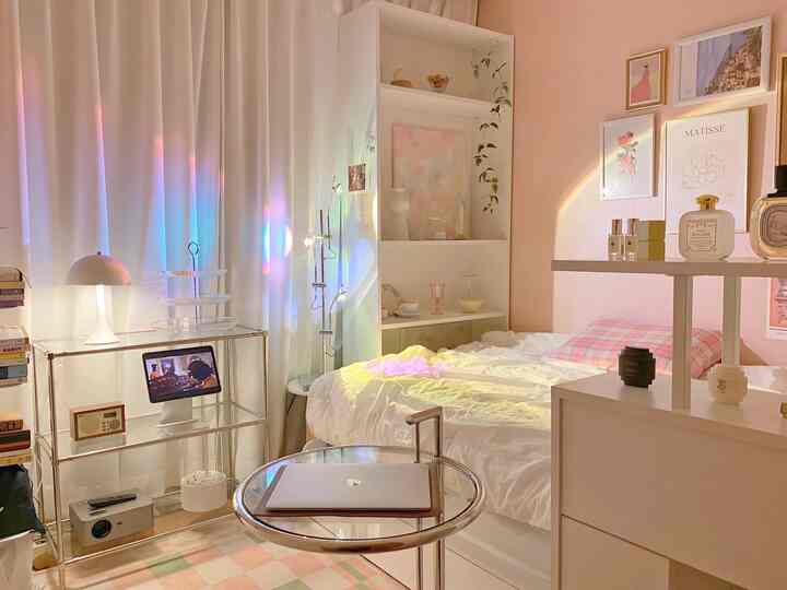 Pastel-toned small bedroom with transparent furniture, white bed and curtains, wall-mounted art frames creating a warm and cozy atmosphere.