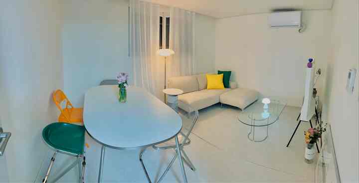 White-toned living room featuring beige sofa, colorful folding chairs, glass coffee table, and floor lamp in a simple modern setting
