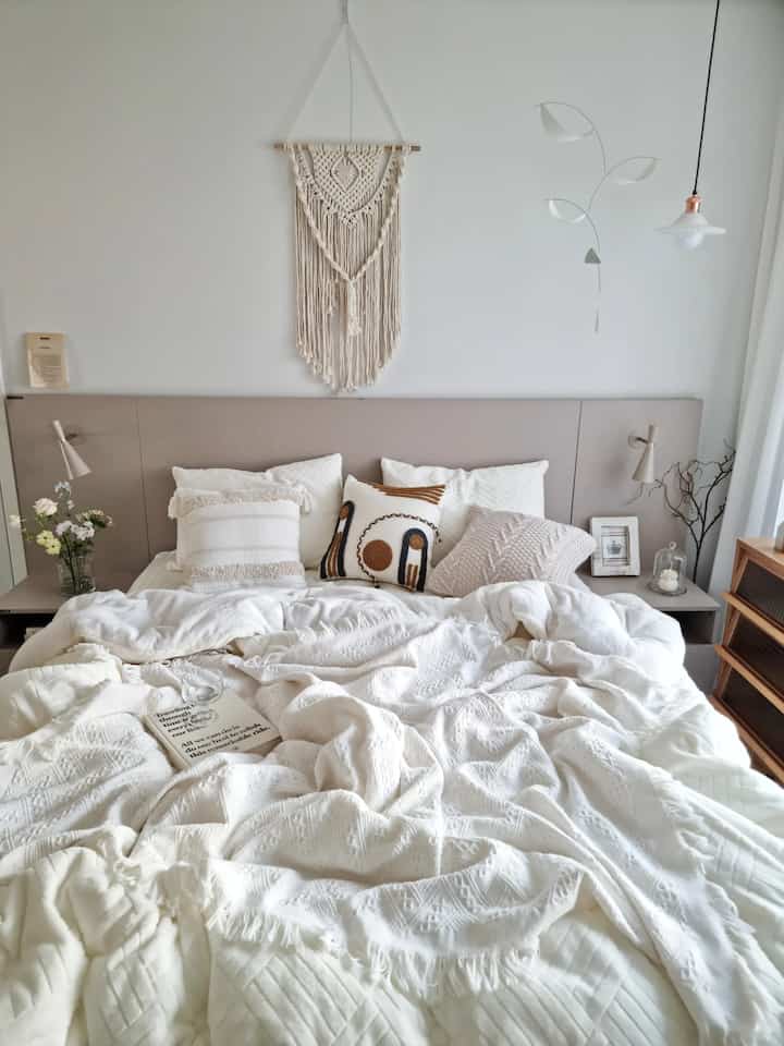 ホワイトとベージュの調和が美しい寝室、心地よいベッドとボヘミアンマクラメが配置された雰囲気のあるナチュラルモダンなインテリア