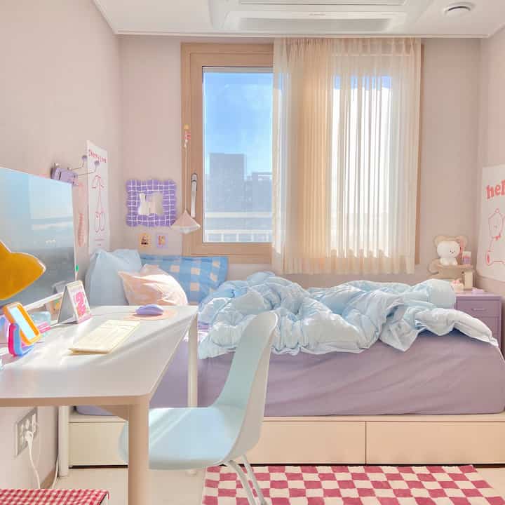 柔らかいベージュ色の壁と青い寝具がある一人暮らしの寝室、窓際に机と椅子が配置された温かみのある空間