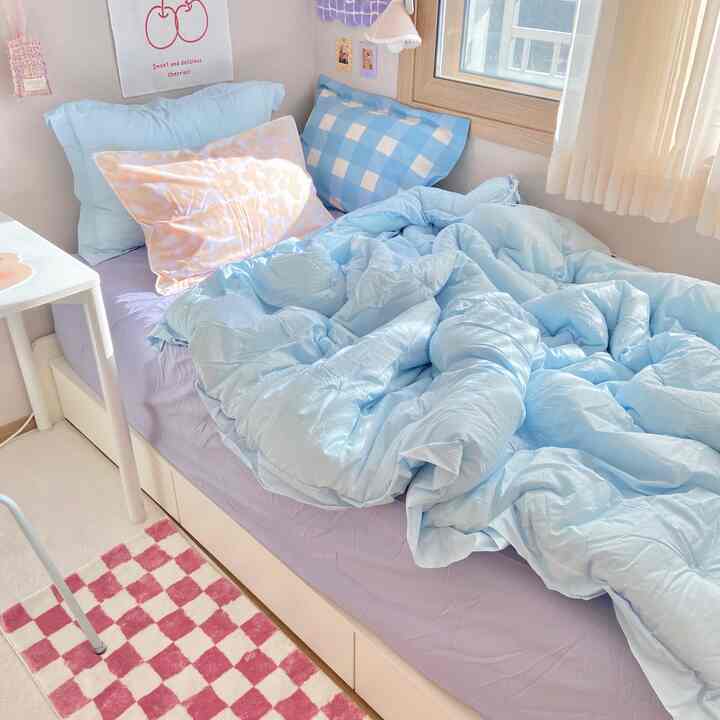 パステル調の寝室、淡いスカイブルーの寝具とピンクのチェック柄ラグが配置されたミニマルで居心地の良い空間