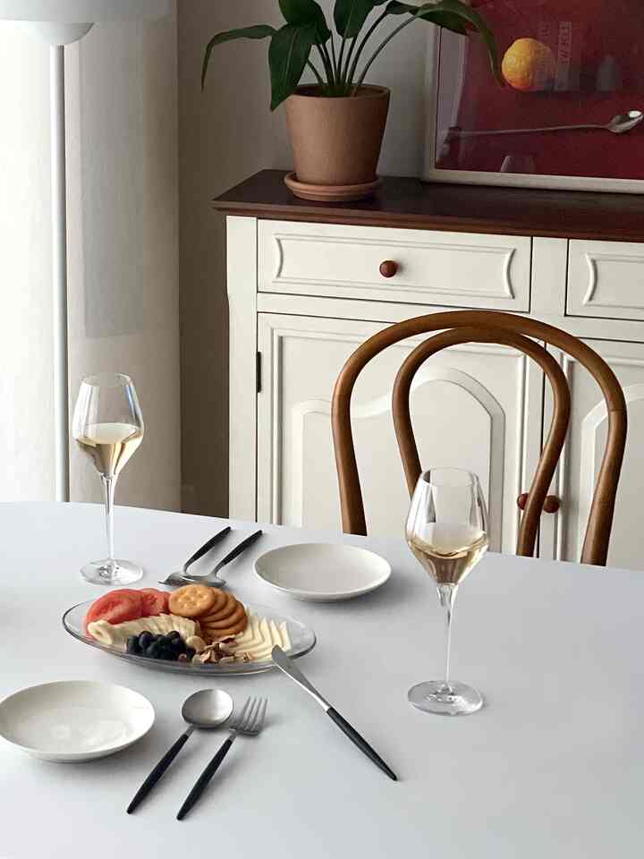 ナチュラルな色調のダイニング、白いダイニングテーブルと木製の椅子、ワイングラスと料理が配置された雰囲気のあるインテリア