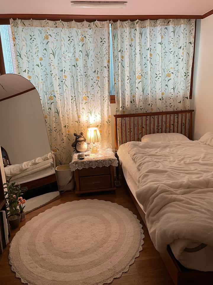 Cozy bedroom with warm wood tones and floral patterned curtains, featuring a wooden bed frame, lace-covered nightstand, and round rug