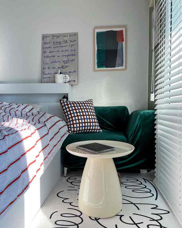 Modern bedroom in white and green tones featuring striped bedding, green sofa, and white blinds by the window