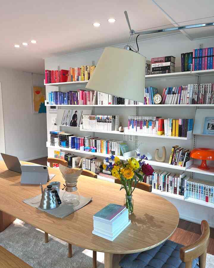 白い壁の棚と木目調のダイニングテーブルが配置された書斎兼リビング、書籍と花が暖かい雰囲気を醸し出す空間