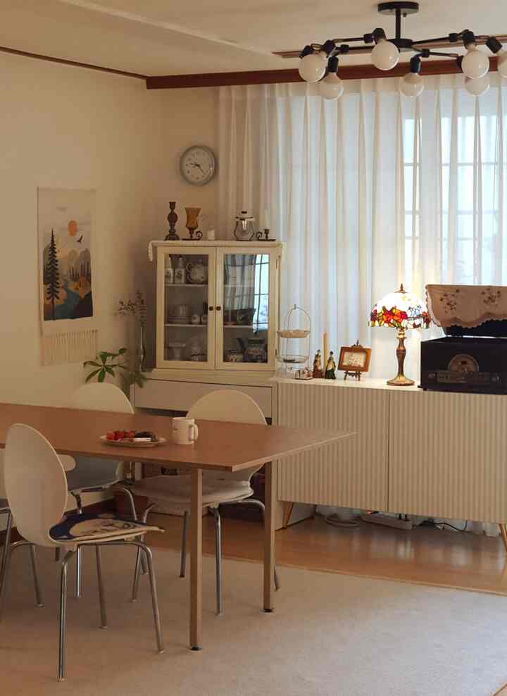 A white and wood-toned dining room featuring curtains, a cabinet, and a stained glass table lamp with a cozy aesthetic