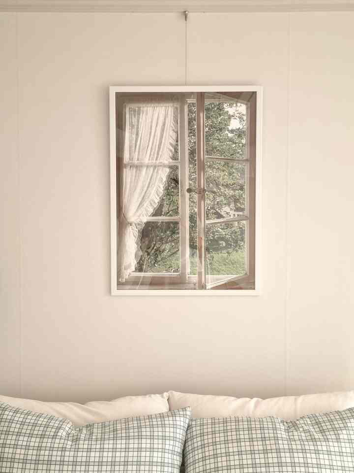 ホワイト調の寝室の壁に窓と緑の風景が描かれたポスター額が掛かり、チェック柄のクッションが置かれた温かみのある空間