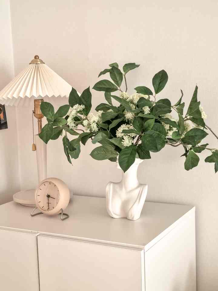 A natural and minimal bedroom nightstand in white and green tones featuring a vase with artificial flowers, clock, and a lamp.