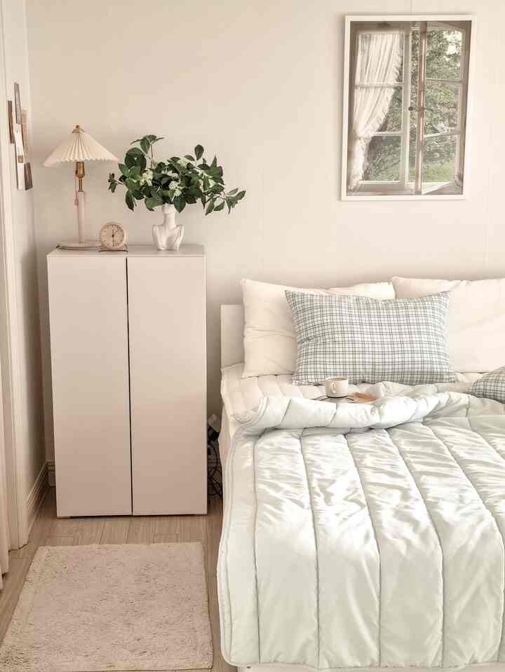 Soft white and beige bedroom featuring a tidy bed, nightstand, plant, and lamp creating a cozy natural atmosphere
