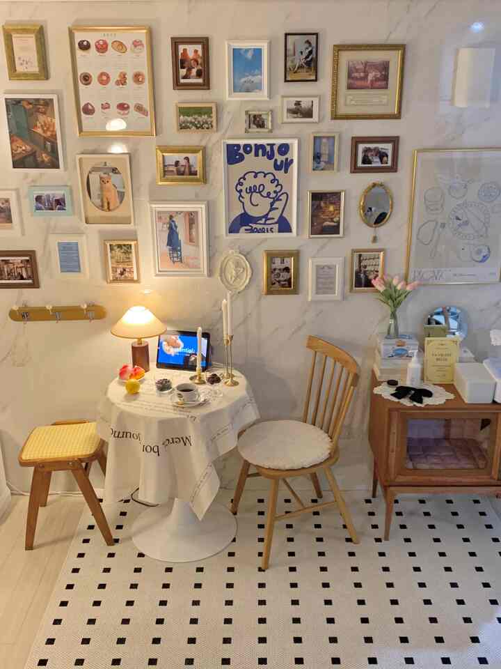 ナチュラルな木目調とゴールドの額縁が調和する小さなカフェ風の部屋、壁一面にアートが飾られ、テーブルにホームカフェのセットが配された雰囲気のあるインテリア