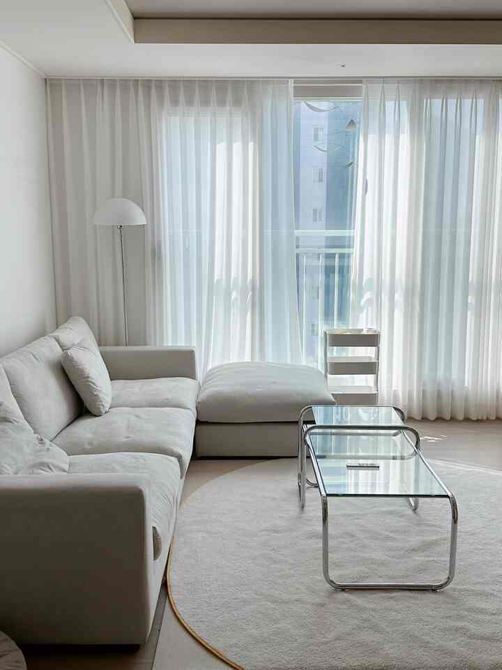 A white and natural tone living room featuring a large sectional sofa, transparent glass coffee table, wide window with sheer curtains, showcasing a simple and modern interior