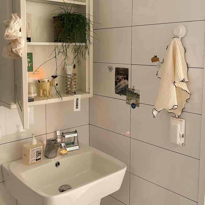 白いタイル壁と洗面台があるアットホームなバスルーム、壁掛け収納、クリーム色のタオル、小さな植物が配置された清潔感ある空間
