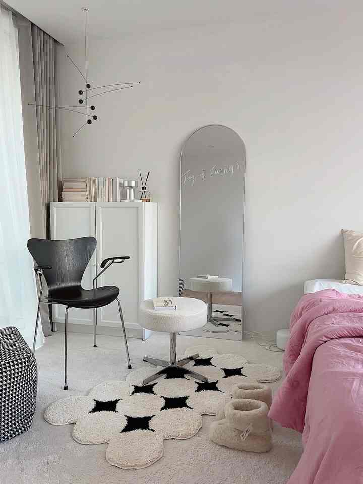 A bedroom with white and black tones accented by pink bedding, featuring an arched full-length mirror and stool in a simple, natural style