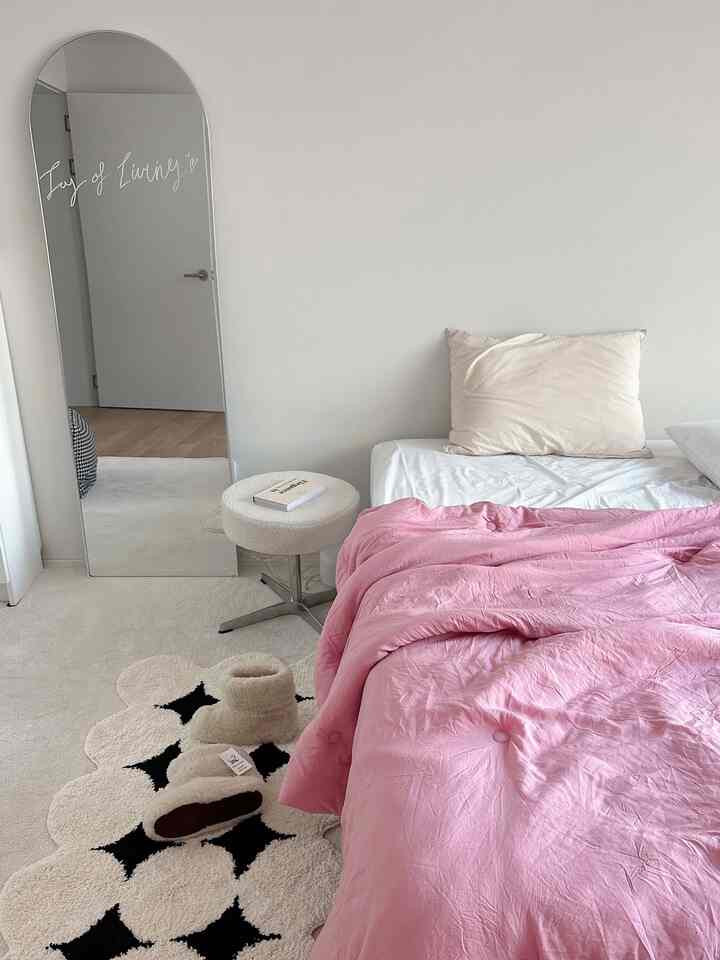 White and pink toned bedroom featuring pink bedding, an arch-shaped full-length mirror, and a floral rug in a bright, clean atmosphere