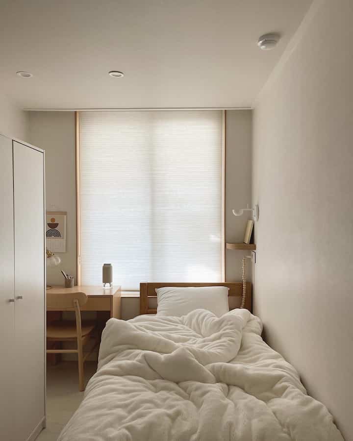 明るい木目調とホワイトトーンが調和したミニマルな一人暮らし用寝室、ふんわりした極細マイクロファイバーの布団と木製ベッドフレームが配置された落ち着いた空間