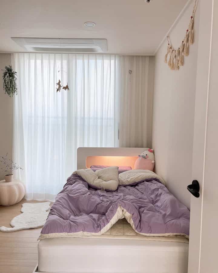 Bright white and lavender toned kids' room featuring sheer curtains and tidy bed with soft natural decor