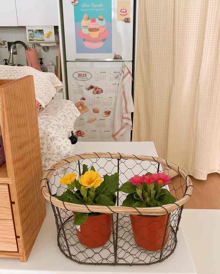 Natural kitchen space with beige curtains and white table featuring wire basket with wooden trim holding yellow and pink potted flowers centered