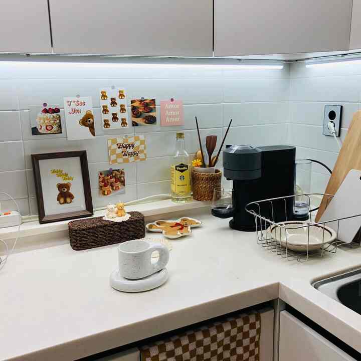 白いタイルの壁とベージュのキッチンカウンター、コーヒーマシンと額縁が配置されたシンプルで清潔感のあるおうちカフェのキッチンスペース