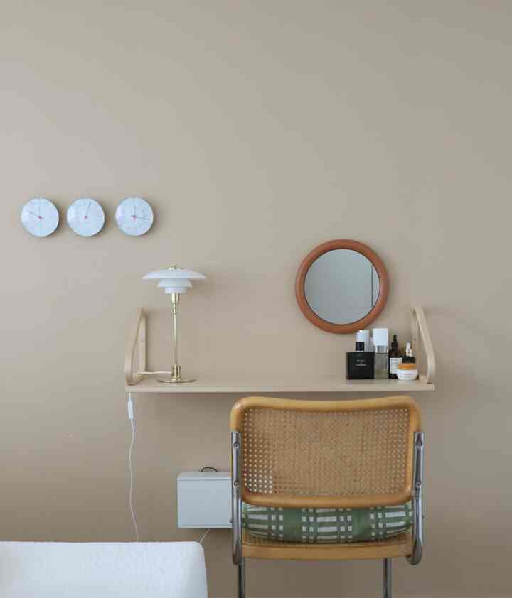 ベージュ色調の寝室の壁に配置されたアルテックの棚、丸い鏡、テーブルランプ、ラタンチェアがある雰囲気のある化粧台スペース