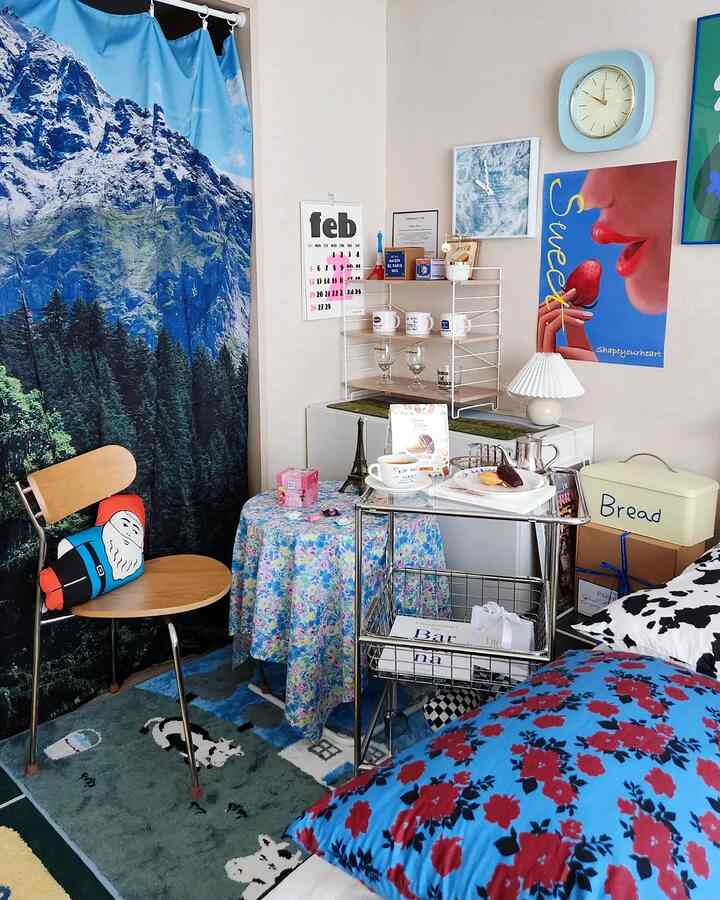 A blue and green toned bedroom featuring mountain scenic curtains, floral bedding, decorative accessories, and a wall calendar creating a cozy space