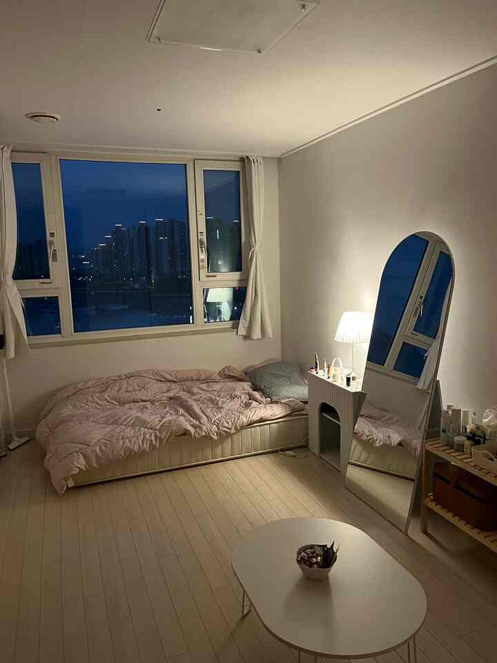 白い壁と木目調の床が調和するワンルーム、ミニマルなマットレス配置と照明、全身鏡が置かれた雰囲気のある空間