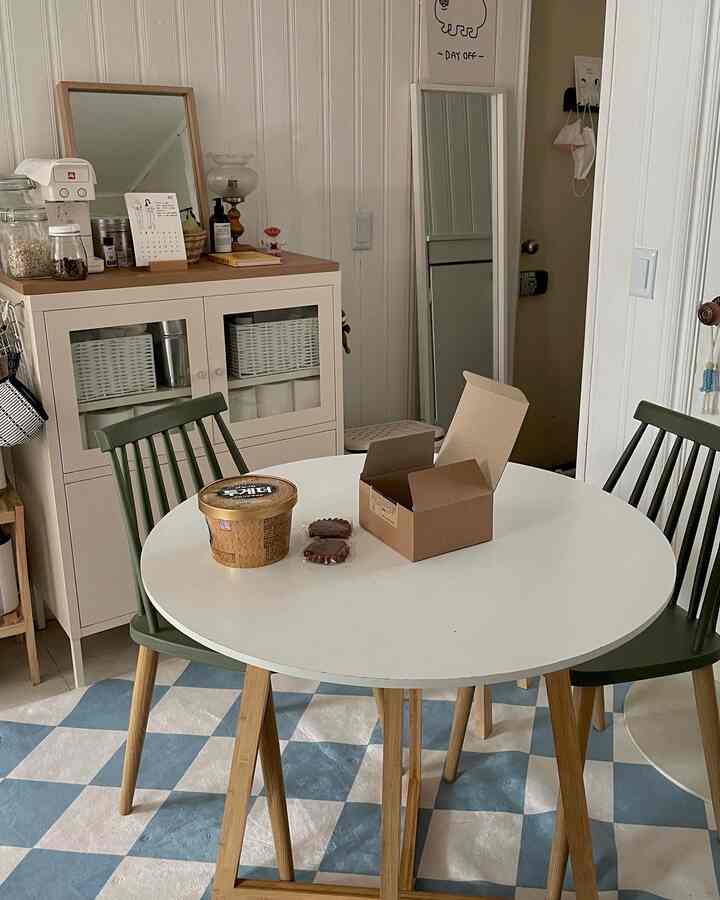 A white and wood tone kitchen dining space featuring a round table, two green chairs, and a beige cabinet with a modern minimalist feel