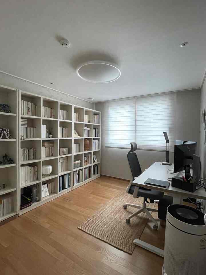 A clean home office study with white and wood tones, featuring a large bookshelf, integrated desk, and an office chair