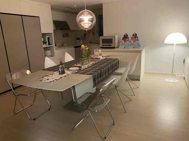 ホワイトとナチュラルカラーのキッチン兼ダイニングルーム、拡張型ダイニングテーブルと白および透明の椅子が配置されたモダンな空間