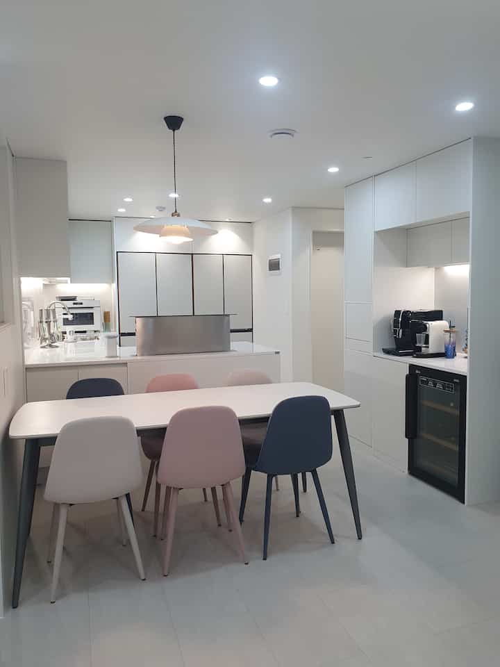 Bright white tone narrow kitchen featuring multicolored dining chairs and pendant light, creating a clean atmosphere