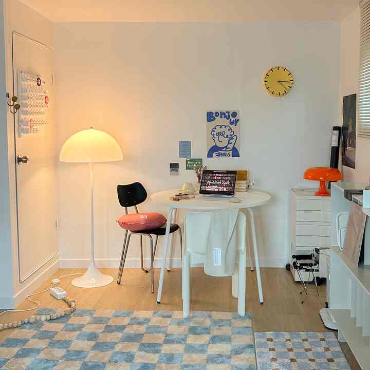 白い壁と木目調の床が特徴の小さなダイニングルーム、丸テーブルとシンプルな椅子、フロアランプとオレンジ色のテーブルランプが調和した雰囲気のあるインテリア