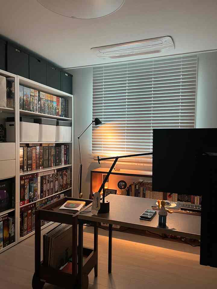 ナチュラルカラーと木目調の調和が取れた書斎、広い本棚とデスク、ブラインドが配置された雰囲気のある趣味の空間