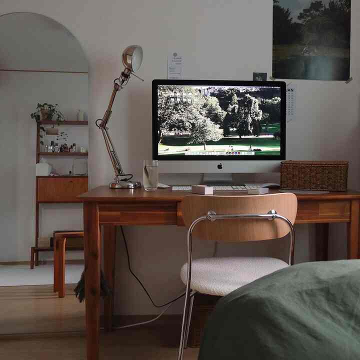 ナチュラルなトーンのワンルームのホームオフィス、木製の机と椅子、大きなiMac、クロームの作業灯が配置された雰囲気のある空間