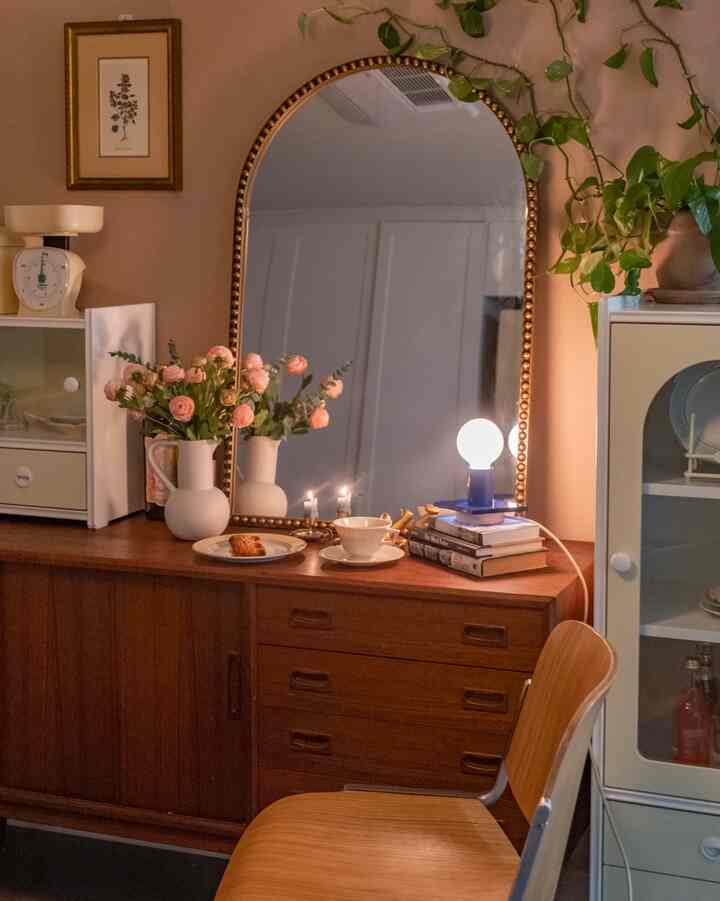 暖かいナチュラルカラーのヴィンテージスタイルの空間、木製キャビネットと金縁のアーチ型鏡、照明と植物が配置された雰囲気のあるインテリア