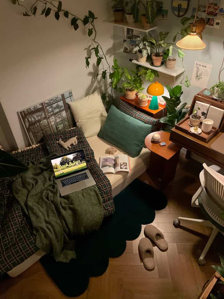 グリーンとブラウンの色調のワンルーム寝室、ベッドやデスク、たくさんの植物が温かい照明の中に配置された雰囲気のあるインテリア