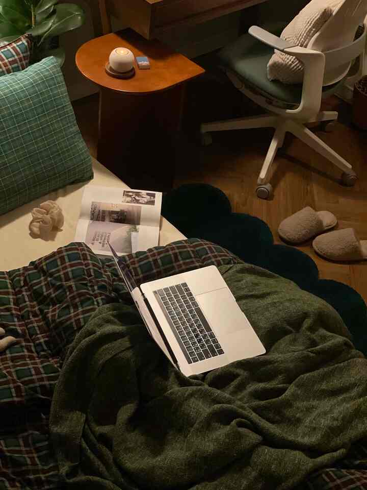 A cozy bedroom space in green and beige tones featuring a laptop, bedding, and home office setup with a warm natural atmosphere