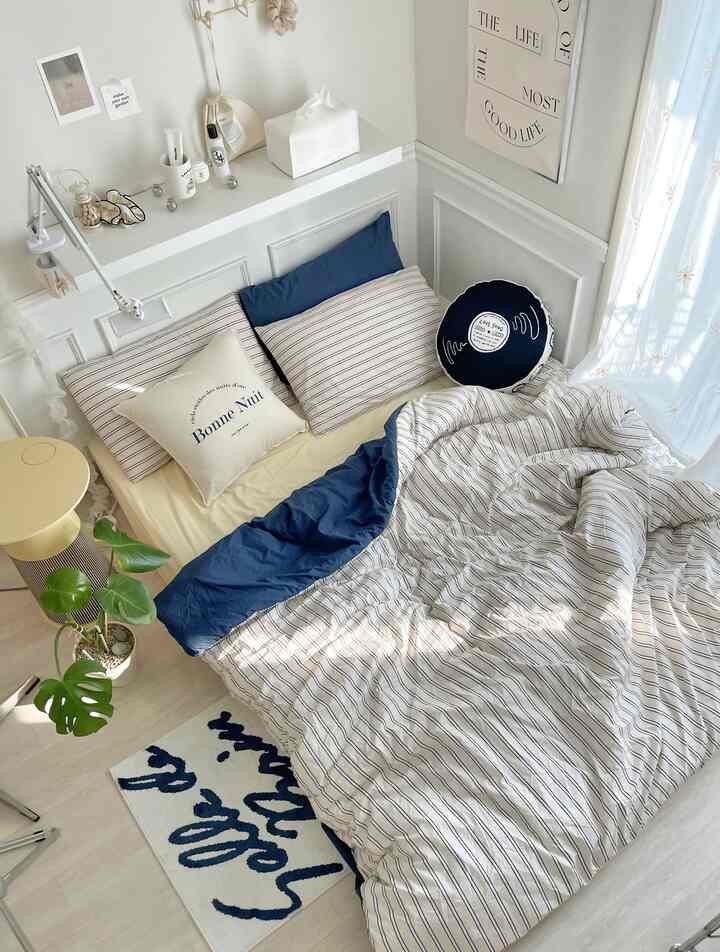 ホワイトとブルーを基調としたワンルームの寝室、ブルーストライプの寝具と多彩なクッションが配された居心地の良い空間
