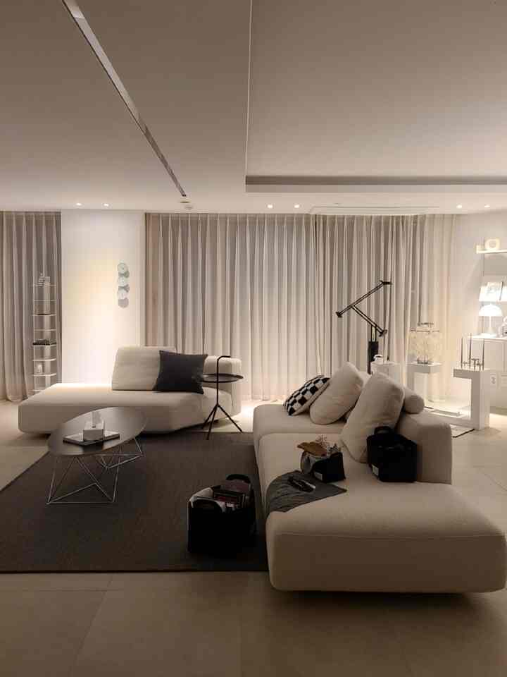 Modern white-toned living room featuring cream sofas, dark brown rug, and soft blackout curtains creating a cozy atmosphere